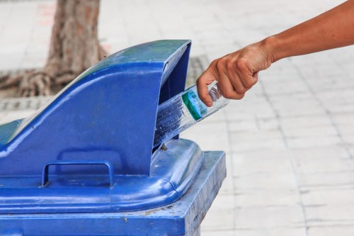 A person’s hand is shown inserting a plastic water bottle into the open top of a blue wheelie bin, which is positioned on a paved sidewalk surface. The bin features a smooth, glossy finish and a curved lid that is partially lifted, revealing its interior designed for waste collection. In the background, a tree trunk and a paved area are visible, suggesting an outdoor urban or residential setting. The scene is well-lit with natural daylight, emphasizing the vibrant blue colour of the bin and the transparency of the water bottle. This image relates to rubbish removal services, illustrating proper disposal of recyclable items typically handled by companies such as Office Clearance Blackwall in the London area, near postcode E14. The focus remains on the action of waste disposal within an outdoor environment suitable for waste collection and management services in the Blackwall locality.