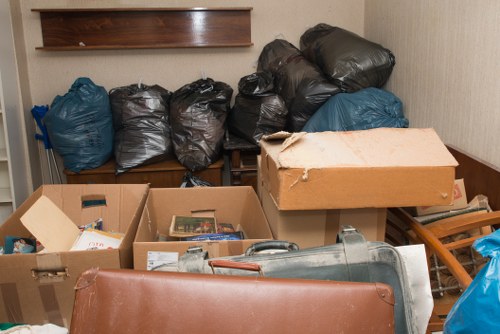 A collection of black and blue plastic rubbish bags, filled and tied at the top, are stacked and placed on a wooden surface in an indoor setting, with additional cardboard boxes filled with miscellaneous waste items positioned in the foreground. The cardboard boxes contain various materials such as paper and other debris, some of which are partially spilling out. To the right, a rolled-up piece of grey plastic or fabric and a wooden stool are visible, with a beige wall and a wooden shelf in the background. The environment appears to be a cluttered storage or disposal area, potentially associated with office clearance services offered by Office Clearance Blackwall in the vicinity of Blackwall, London. The scene emphasizes waste collection and removal, relevant to rubbish clearance tasks typically handled by a professional rubbish removal service.