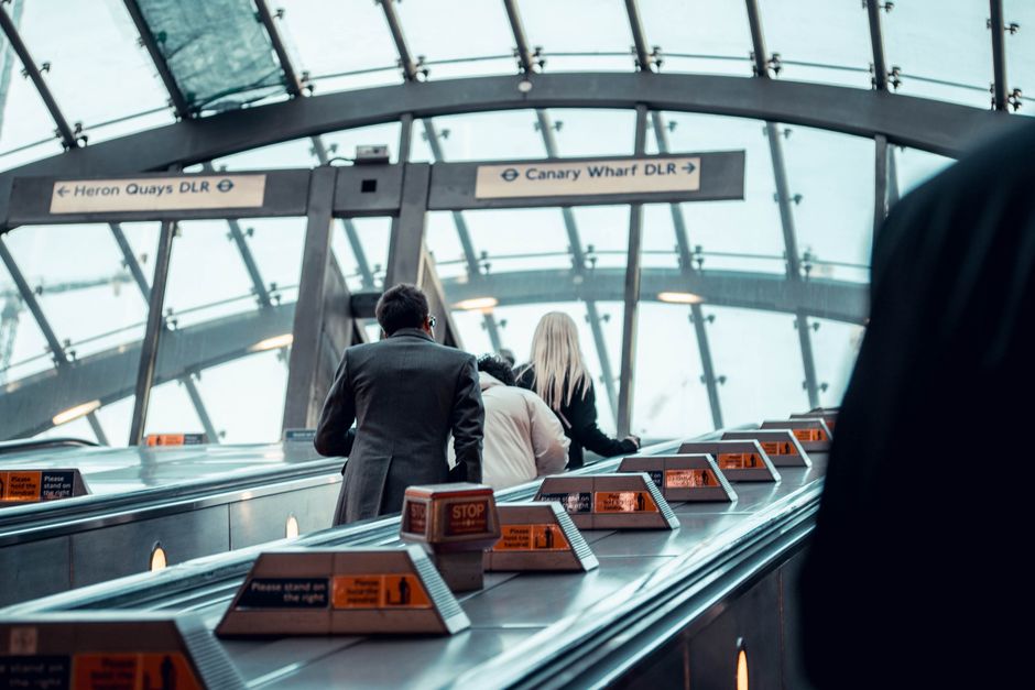 A worker wearing a grey beanie, black jacket, and white gloves stands on an outdoor train station platform next to a large metal trolley filled with assorted waste materials, including black, orange,
