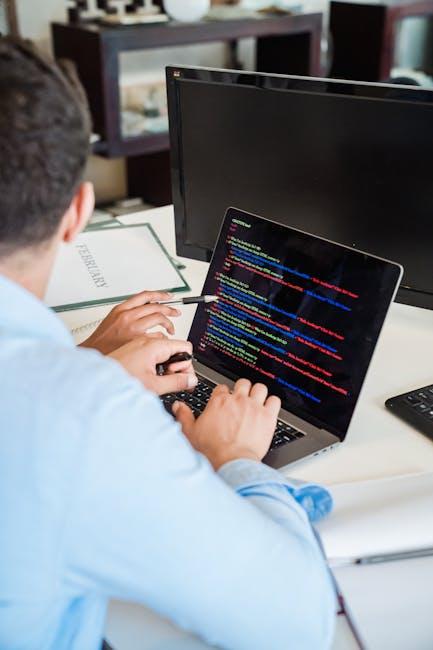 The image depicts a partially visible black laptop with a red-backlit keyboard placed on a dark surface, with the screen displaying lines of blurred code or text in blue, red, and white colors. Next t
